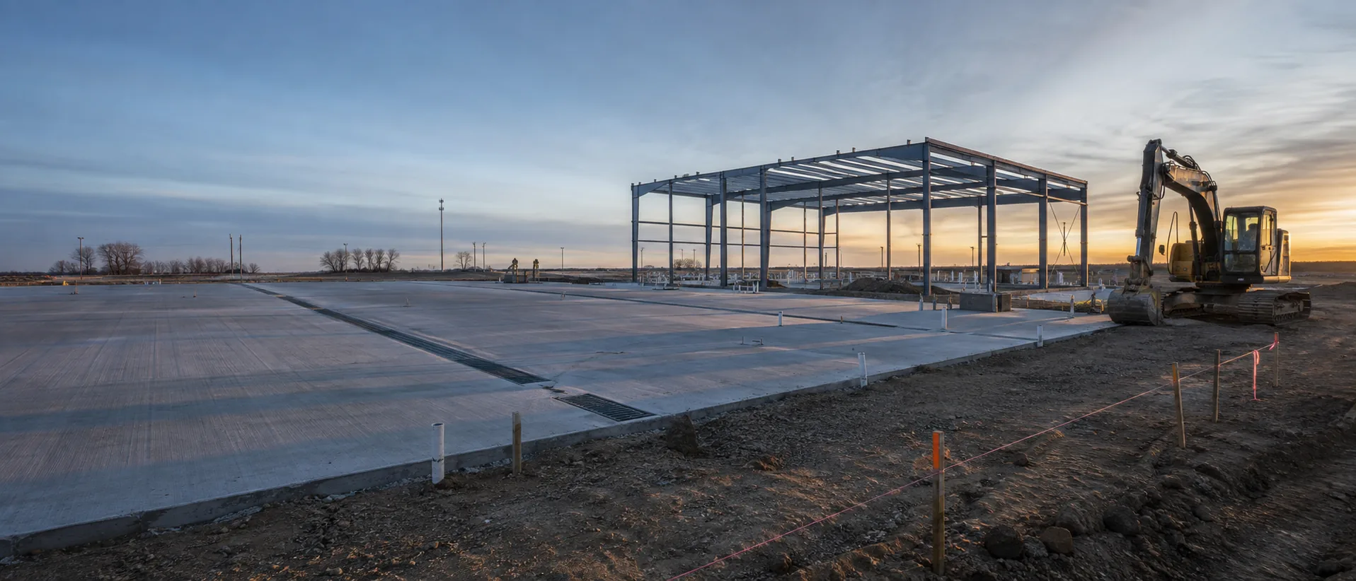 A commercial truck wash under construction at sunrise: a poured concrete slab with drain trenches and a partial steel frame.